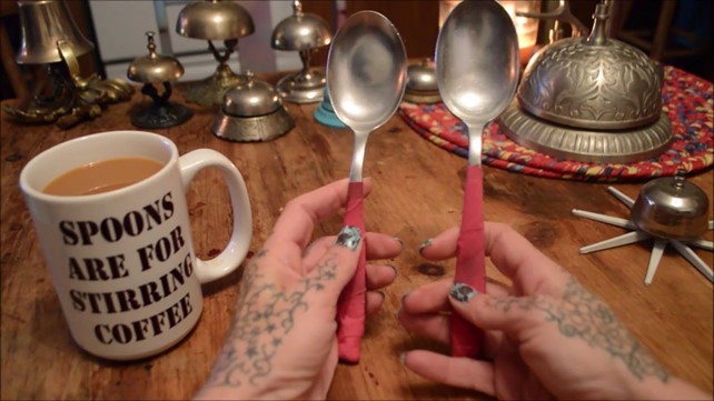 Tattooed hands hold two spoons with pink wrapped handles. Next to them on the table is a full mug of tea with the slogan "SPOONS ARE FOR STIRRING". The background features a number of bells. 
